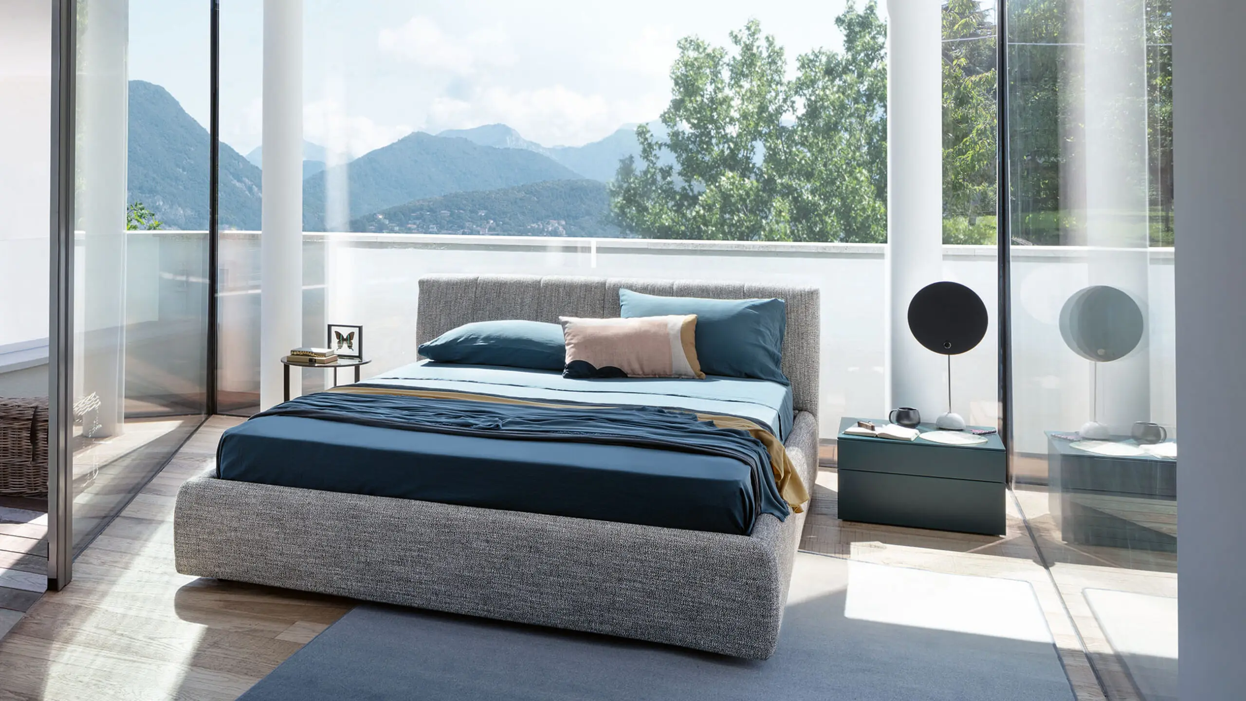 Camera da letto con letto imbottito in tessuto grigio, lenzuola blu e vetrate affacciate su montagne e alberi